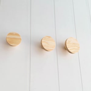 Può includere: Tre ganci da parete rotondi in legno montati su una parete bianca. I ganci sono in legno naturale di colore marrone chiaro.