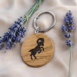 May include: A wooden keychain with a silver ring. The round, light brown wooden pendant features a black silhouette of a ram. Lavender sprigs with purple flowers are arranged around the keychain.