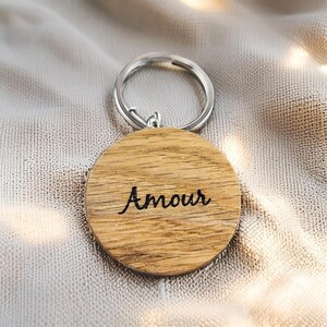 May include: A round wooden keychain with the word "Amour" in cursive script. The keychain has a silver metal ring and a light brown wooden pendant. The wood grain is visible on the pendant.