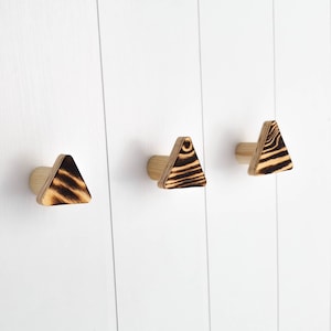 May include: Three wooden triangle hooks with a burnt wood finish mounted on a white wall.