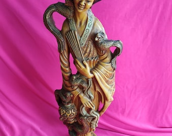 May include: A carved wooden figurine of a woman in traditional Chinese clothing holding a fan and a bird. The figurine is painted in shades of brown and gold and stands on a decorative base.