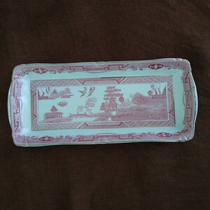 Puede incluir: Una bandeja rectangular para servir con fondo blanco y borde rosa y dorado. La bandeja presenta un diseño chino tradicional con una pagoda, árboles y pájaros.