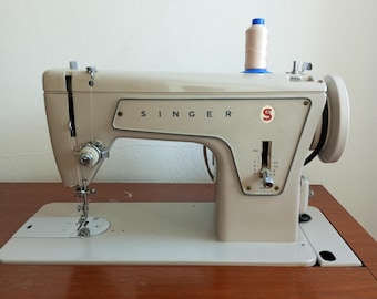 May include: A beige Singer sewing machine featuring a horizontal spool pin with a beige thread spool, a vertical stitch length adjustment lever, a needle bar, a presser foot, and a handwheel on the right side. The machine body displays the Singer logo.