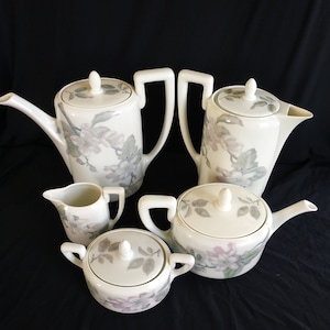 Könnte beinhalten: Ein Set aus fünf Porzellan-Teetassen und einer Teekanne mit Blumenmuster. Die Tassen und die Teekanne sind weiß mit einem zarten Blumenmuster in Rosa- und Grüntönen.