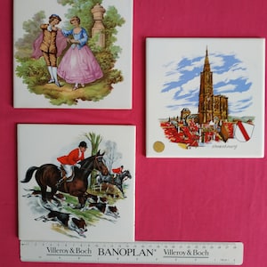 May include: Three vintage ceramic tiles with colorful illustrations. The first tile features a couple in a garden setting. The second tile depicts a cathedral in Strasbourg, France. The third tile shows a hunter on horseback with hounds chasing a fox.