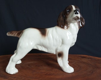 Peut inclure: Une figurine en céramique blanche et brune d'un chien Springer Spaniel. Le chien est debout, la tête inclinée sur le côté et les oreilles dressées.