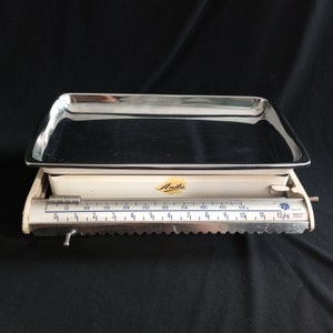 May include: A white kitchen scale with a stainless steel weighing tray. The scale has a black and white ruler with markings up to 12 kg. The scale is labeled "Anila" in gold lettering.
