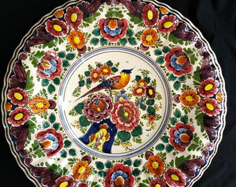 Könnte beinhalten: Ein weißer Keramikteller mit blau-weißem Rand und einem farbenfrohen Blumendesign. In der Mitte des Tellers befindet sich ein Vogel mit einem rot-orange-gelben Design.