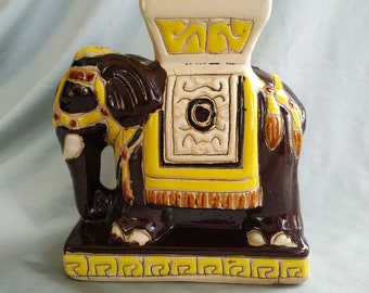 May include: A ceramic elephant figurine featuring a dark brown glossy glaze with bright yellow and cream-colored decorative accents. The elephant is adorned with a saddle-like structure on its back and stands on a rectangular base with a yellow pattern.