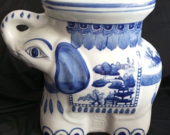 May include: A white ceramic elephant stool with blue and white painted designs. The stool features a traditional Asian design with a pagoda, trees, and mountains. The elephant's trunk is curled up and its eyes are painted blue.