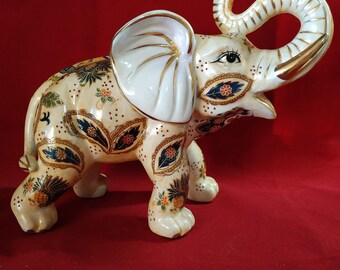 May include: A white ceramic elephant figurine with gold and blue painted details. The elephant has a raised trunk and is decorated with a paisley pattern.
