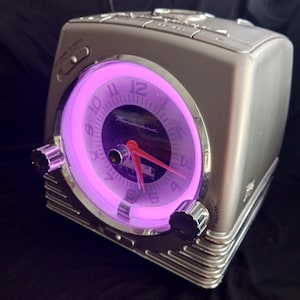 May include: A silver clock radio with a purple glow around the clock face. The clock face has black numbers and the word "Thunderbird" printed on it. The radio has a volume knob and other controls.