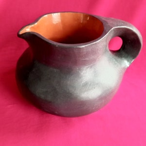 May include: A black ceramic pitcher with a rounded body and a handle. The pitcher has a terracotta colored interior.