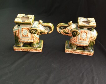 May include: Two green and brown ceramic elephant candle holders with intricate details. The elephants are facing each other and have a raised platform on their backs.