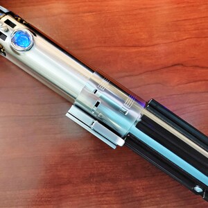 Puede incluir: Un sable de luz plateado con un mango negro y una hoja de sable de luz azul. El sable de luz est&aacute; sobre una superficie de madera.