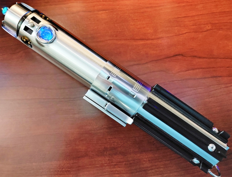 Puede incluir: Un sable de luz plateado con un mango negro y una hoja de sable de luz azul. El sable de luz est&aacute; sobre una superficie de madera.