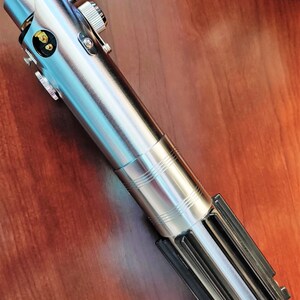 Puede incluir: Un sable de luz plateado con un mango negro y una hoja plateada. El sable de luz tiene un dise&ntilde;o detallado y est&aacute; hecho de metal.