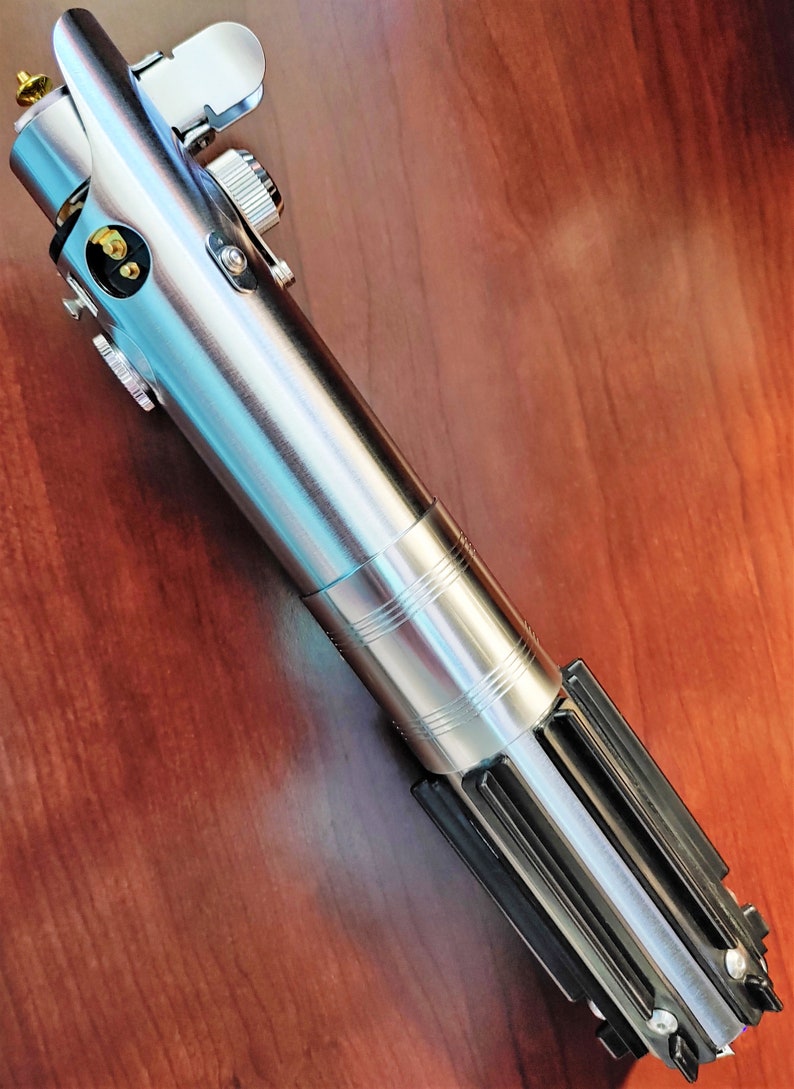 Puede incluir: Un sable de luz plateado con un mango negro y una hoja plateada. El sable de luz tiene un dise&ntilde;o detallado y est&aacute; hecho de metal.