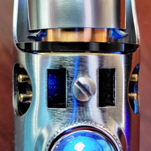 Puede incluir: Una empu&ntilde;adura de sable de luz plateada con una luz LED azul y una parte superior dorada. La empu&ntilde;adura tiene una superficie texturizada y un peque&ntilde;o tornillo en el lateral.