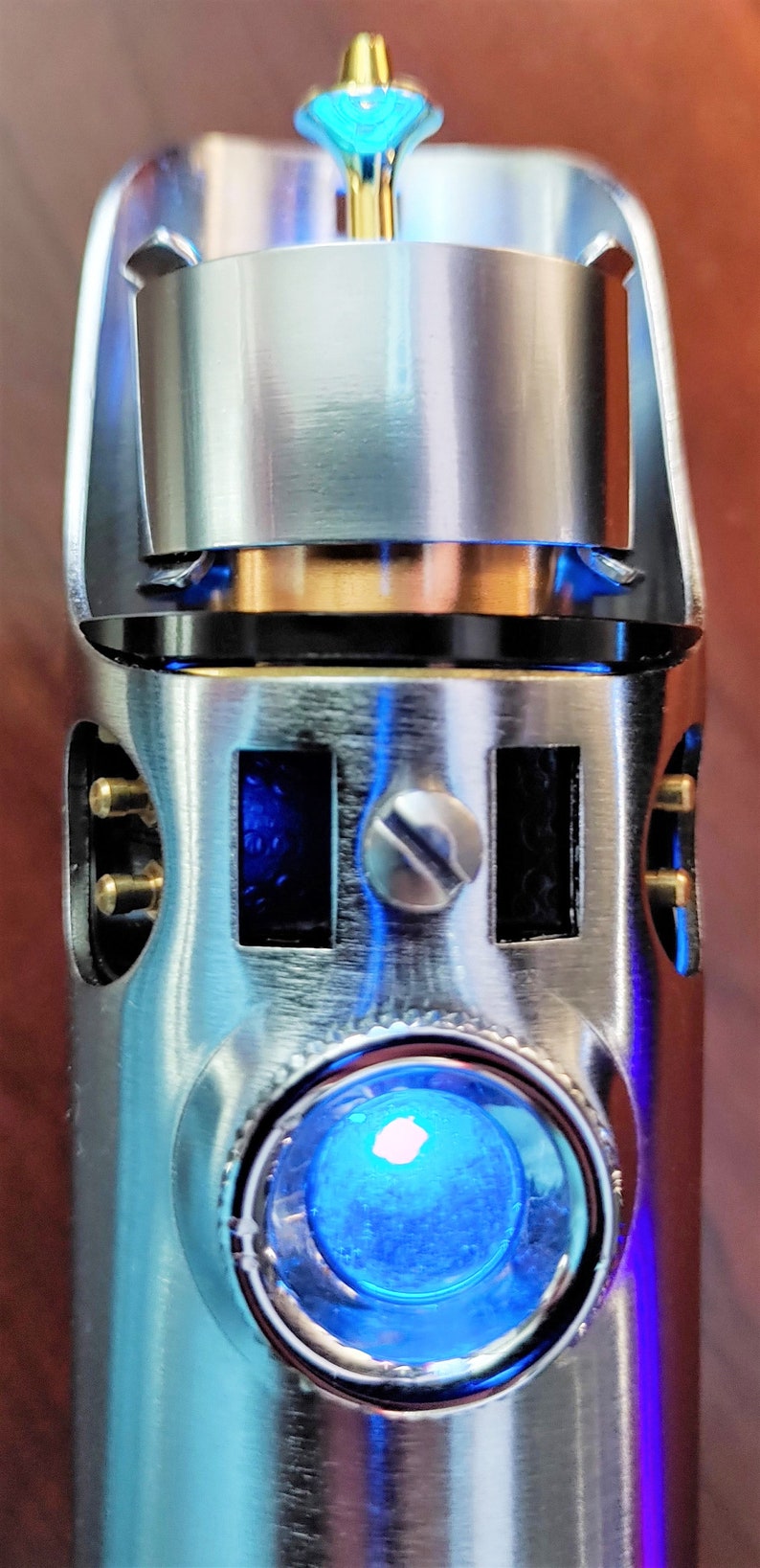 Puede incluir: Una empu&ntilde;adura de sable de luz plateada con una luz LED azul y una parte superior dorada. La empu&ntilde;adura tiene una superficie texturizada y un peque&ntilde;o tornillo en el lateral.