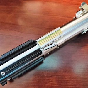 Puede incluir: Un sable de luz plateado y negro con un interruptor de activaci&oacute;n verde y un detalle dorado en el extremo.