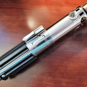 Puede incluir: Un sable de luz plateado y negro con un mango detallado y una hoja plateada. El sable de luz est&aacute; sobre una superficie de madera.