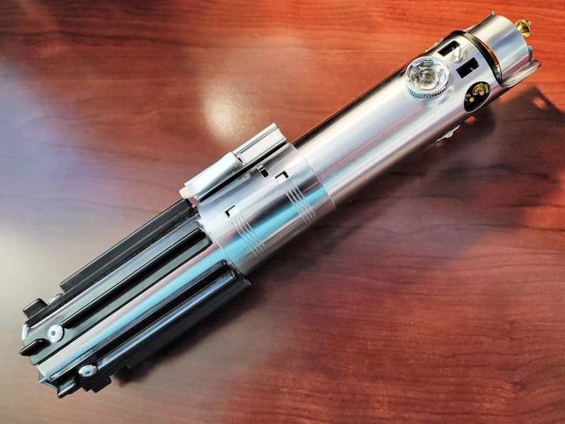 Puede incluir: Un sable de luz plateado y negro con un mango detallado y una hoja plateada. El sable de luz est&aacute; sobre una superficie de madera.