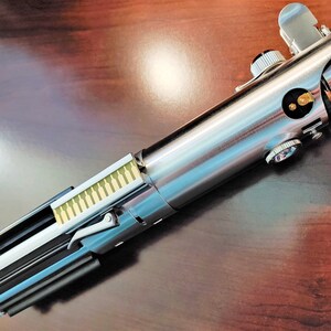 Puede incluir: Un sable de luz plateado y negro con un dise&ntilde;o detallado. El sable de luz tiene un mango plateado con un agarre negro y una hoja plateada. El sable de luz es un objeto de colecci&oacute;n popular para los fan&aacute;ticos de la franquicia Star Wars.