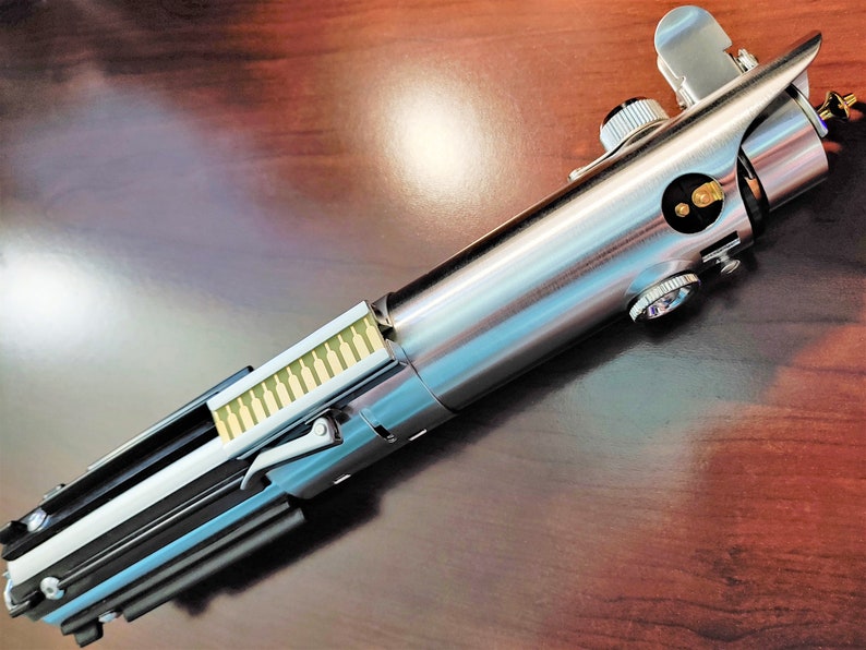 Puede incluir: Un sable de luz plateado y negro con un dise&ntilde;o detallado. El sable de luz tiene un mango plateado con un agarre negro y una hoja plateada. El sable de luz es un objeto de colecci&oacute;n popular para los fan&aacute;ticos de la franquicia Star Wars.