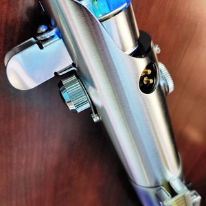 Puede incluir: Una empu&ntilde;adura de sable de luz plateada con una hoja de sable de luz azul. La empu&ntilde;adura tiene un pomo plateado y una empu&ntilde;adura negra. La empu&ntilde;adura est&aacute; hecha de metal y tiene un dise&ntilde;o detallado.