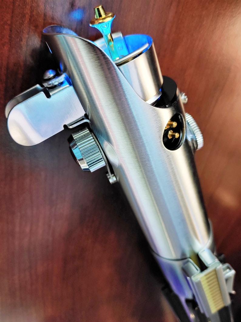 Puede incluir: Una empu&ntilde;adura de sable de luz plateada con una hoja de sable de luz azul. La empu&ntilde;adura tiene un pomo plateado y una empu&ntilde;adura negra. La empu&ntilde;adura est&aacute; hecha de metal y tiene un dise&ntilde;o detallado.