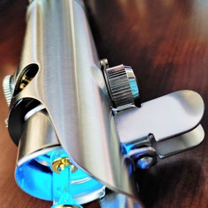 Puede incluir: Un sable de luz plateado con una hoja de sable de luz azul. El sable de luz tiene un mango plateado con un agarre negro y una perilla plateada en el extremo. El sable de luz est&aacute; sobre una superficie de madera.