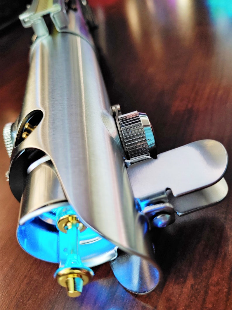 Puede incluir: Un sable de luz plateado con una hoja de sable de luz azul. El sable de luz tiene un mango plateado con un agarre negro y una perilla plateada en el extremo. El sable de luz est&aacute; sobre una superficie de madera.