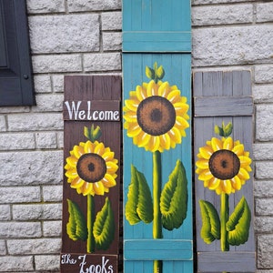 Puede incluir: Tres contraventanas decorativas de madera con girasoles pintados. Las contraventanas son marrones, turquesas y grises. La contraventana marrón tiene las palabras "Welcome" y "The Looks" pintadas. Los girasoles tienen pétalos amarillos, centros marrones y hojas verdes.
