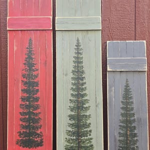 May include: Three wooden decorative shutters in varying sizes and colors. Each shutter features a hand-painted evergreen tree design. The shutters are painted red, green, and gray, with a distressed finish for a rustic look.