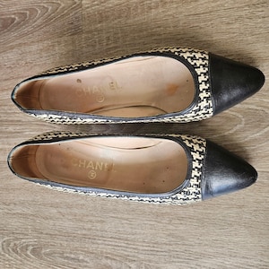 May include: A pair of Chanel pointed-toe flats featuring a woven black and beige pattern across the body, solid black leather toe caps, and black leather trim along the opening. The interior insoles are beige and feature the Chanel brand name.