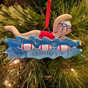 May include: A swimmer ornament with a red ribbon for hanging. The swimmer is in a blue wave with the text "Stone Swim Club", "2020", and "Jennifer's #1". The swimmer wears goggles and a swim cap.