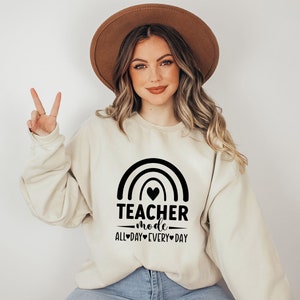 May include: A beige sweatshirt with a black rainbow and the text "Teacher Mode All Day Every Day" with a heart in the rainbow.