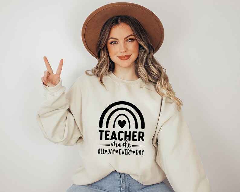 May include: A beige sweatshirt with a black rainbow and the text "Teacher Mode All Day Every Day" with a heart in the rainbow.