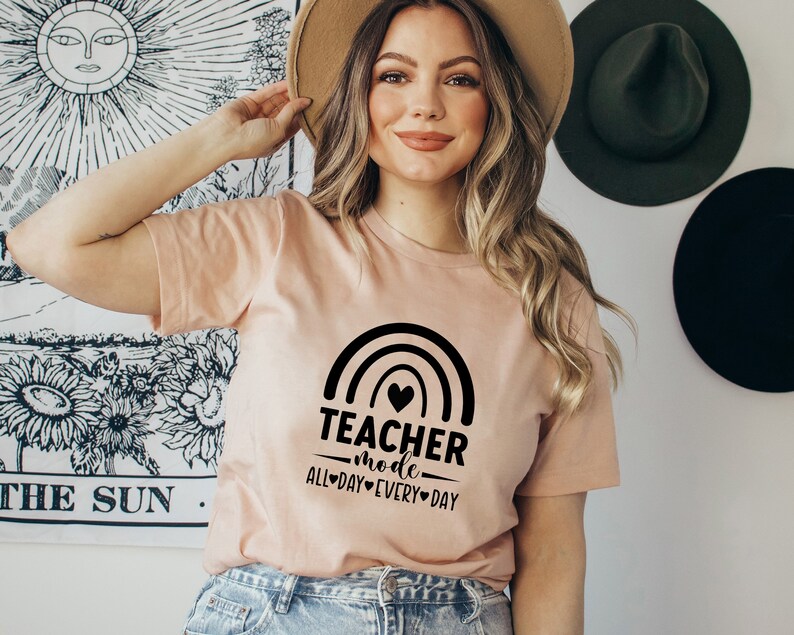 May include: A peach-colored t-shirt with a black rainbow graphic and the text "Teacher mode all day every day".