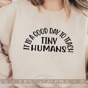May include: A beige sweatshirt with the text "It is a good day to teach tiny humans" in black lettering.