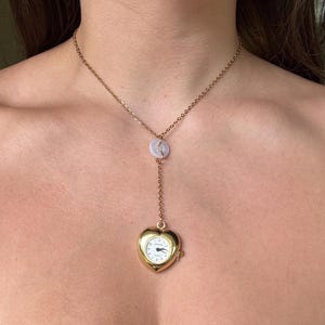 May include: A gold-toned necklace featuring a delicate chain with a small, translucent circular button accent. The pendant is a heart-shaped gold-toned watch case with a white dial, black hands, and black numerals, suspended from the chain in a lariat style.