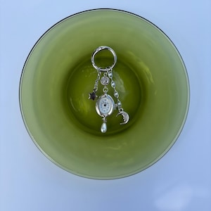 Könnte beinhalten: Ein silberfarbener Schlüsselanhänger mit einem ovalen analogen Zifferblatt mit schwarzen Ziffern. Die Uhr hängt an einem runden Ring zusammen mit baumelnden Anhängern, darunter ein kleiner Stern, ein Halbmond mit Gesicht und eine weiße Perle.