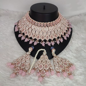 Peut inclure: Un ensemble de bijoux de couleur or rose comprenant un collier et des boucles d'oreilles assorties. Le collier et les boucles d'oreilles sont ornés de cristaux transparents et d'accents roses en forme de goutte, avec des pampilles de perles roses. L'ensemble est présenté sur une tête de mannequin noire.