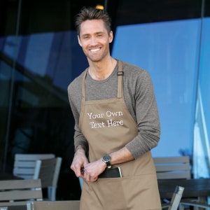 May include: A brown apron with a large pocket and adjustable straps. The apron has white embroidery that says "Your Own Text Here".