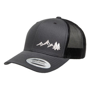 Puede incluir: Una gorra de camionero gris y negra con un diseño de montañas y árboles bordados en blanco. La gorra tiene una parte trasera de malla negra y un cierre a presión. La gorra tiene una etiqueta que dice "The Classics Yupoong Snapback Since 1974".