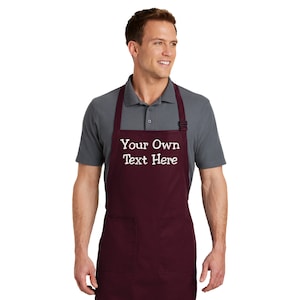 May include: A maroon apron with two pockets and adjustable straps. The apron has white embroidery that says "Your Own Text Here".