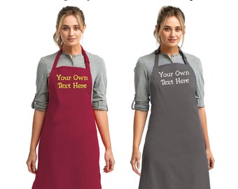 Puede incluir: Dos mujeres con delantales personalizados, uno burdeos y otro gris oscuro, con el texto "Your Own Text Here" bordado en amarillo en el delantal burdeos y en blanco en el delantal gris.