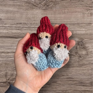 May include: Three small, handmade knitted gnomes. Each gnome has a red hat, a beige face, a fluffy white beard, and a light blue body. The gnomes are held in a hand against a wooden background.