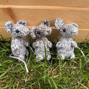 May include: Three handmade gray knitted mice with black eyes and long white tails are displayed on green grass. The mice are standing in front of a wooden background. The knitted toys have large ears and small arms.